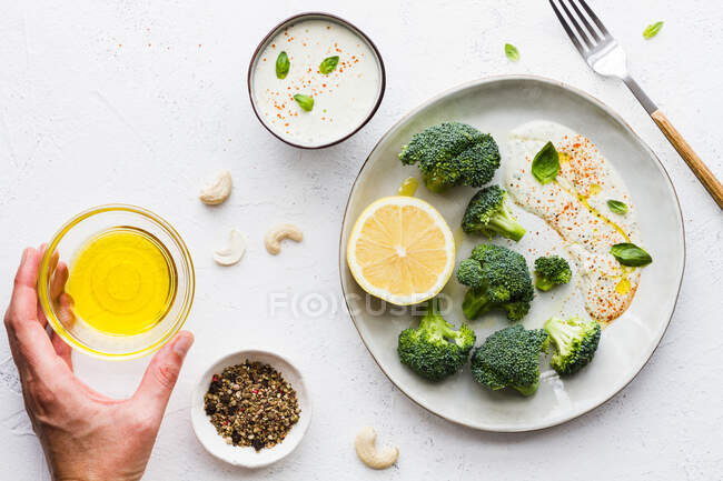 Верхний вид обрезанных неузнаваемых рук, поедающих вкусную брокколи со свежим лимоном и соусом возле миски с оливковым маслом и смесью специй на столе — стоковое фото