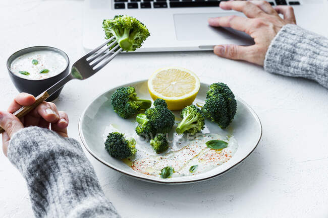 Crop femelle anonyme avec délicieux brocoli cuit à la fourche navigation sur Internet sur netbook à table — Photo de stock