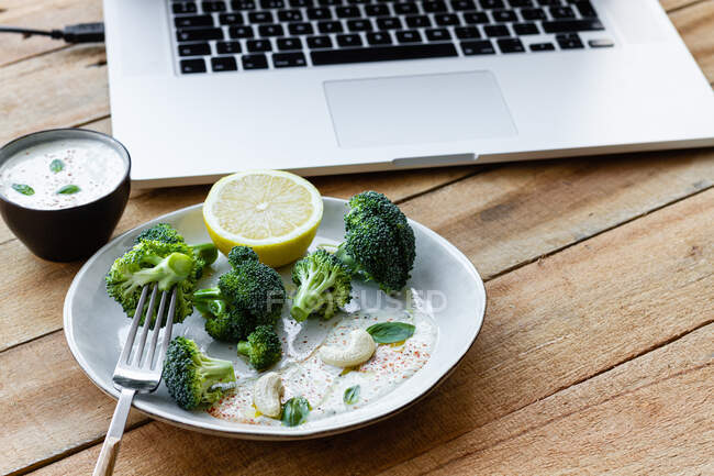 Deliziosi broccoli cotti con fetta di limone e anacardi vicino alla ciotola con salsa bianca e netbook su tavolo di legno — Foto stock
