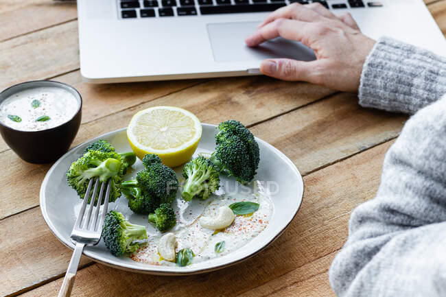 Обрізати анонімну жінку зі смачними вареними броколі на виделці, переглядаючи Інтернет на нетбуці за столом — стокове фото