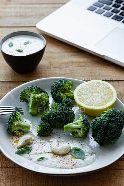 Yummy cooked broccoli with lemon slice and cashew nuts near bowl with white sauce and netbook on wooden table — Stock Photo