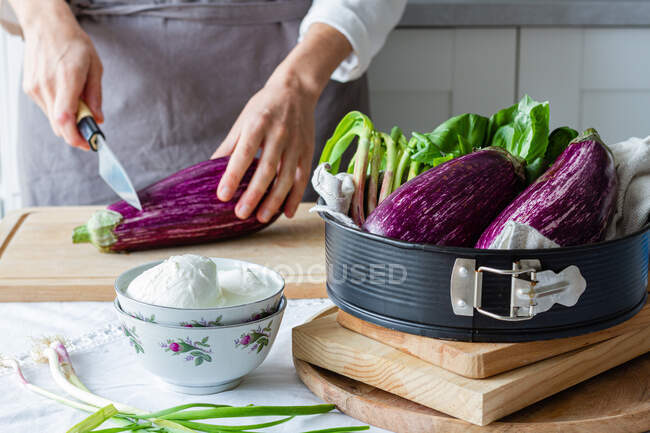 Chef irreconhecível em avental berinjela de corte com faca na placa de corte enquanto cozinha almoço saudável na cozinha — Fotografia de Stock