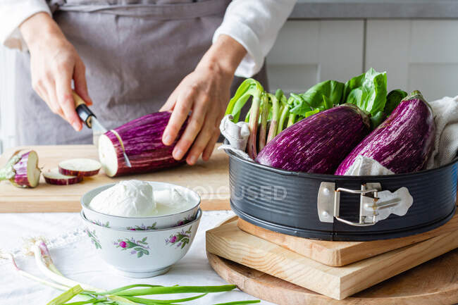 Chef irreconhecível em avental berinjela de corte com faca na placa de corte enquanto cozinha almoço saudável na cozinha — Fotografia de Stock