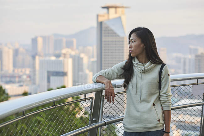Giovane donna asiatica pensierosa in abiti casual in piedi vicino ringhiera e guardando lontano nella moderna città urbana alla luce del giorno — Foto stock