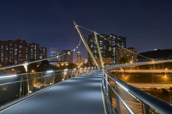 Puente peatonal colgante contemporáneo en diseño creativo cruzando carretera en la ciudad urbana moderna por la noche - foto de stock
