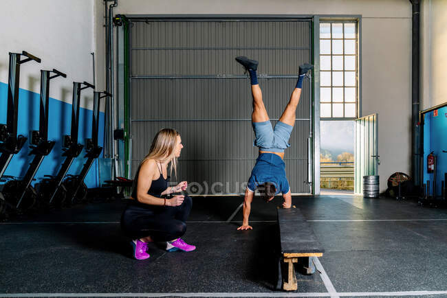 Молода усміхнена жіноча підготовка спортсменки та допомога анонімному спортсмену, що стоїть на руках під час функціональної підготовки в тренажерному залі — стокове фото