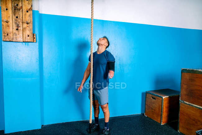 Disabled male athlete in sportswear looking up near workout rope and blue wall in gymnasium — Stock Photo