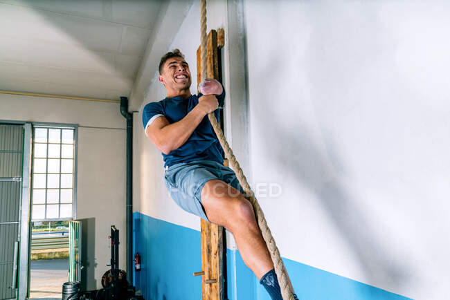 Vista lateral del atleta masculino discapacitado en ropa deportiva escalada cuerda de entrenamiento cerca de la pared brillante en el gimnasio - foto de stock