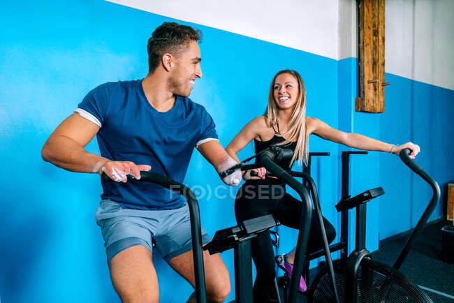 Веселый молодой спортсмен-инвалид и спортсменка катаются на стационарных велосипедах, глядя друг на друга во время функциональной тренировки в тренажерном зале — стоковое фото