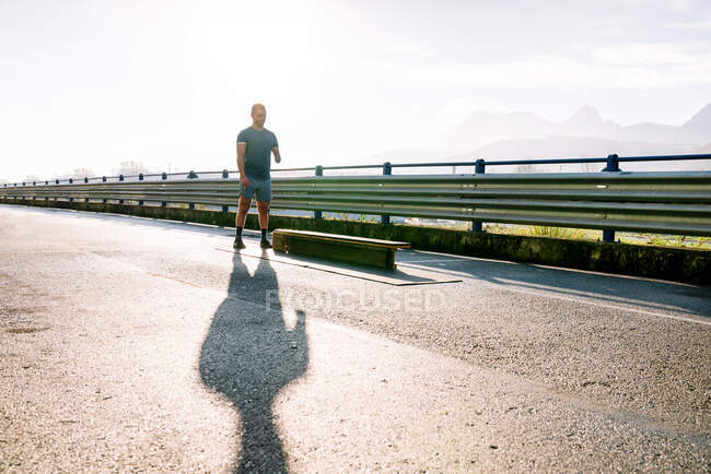Disabled male athlete in sportswear preparing for exercise near bench on fenced bridge with shadow under cloudy sky — Stock Photo