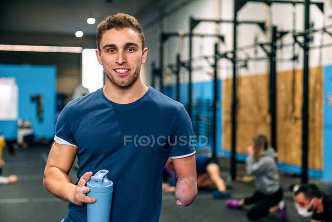 Jovens incapacitados atleta masculino feliz em sportswear segurando garrafa de água enquanto olha para a câmera durante o treino perto de parceiros irreconhecíveis no ginásio — Fotografia de Stock