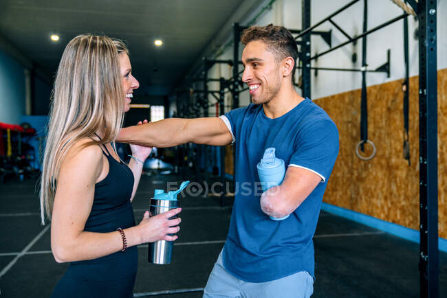 Side view of young sportswoman near handicapped male athlete talking while having a water brake holding bottles while looking at each other during workout near barbells in gym — Stock Photo