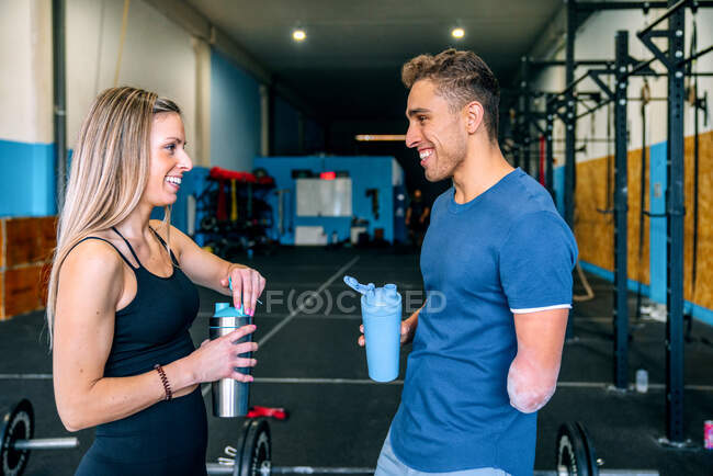 Side view of young sportswoman near handicapped male athlete talking while having a water brake holding bottles while looking at each other during workout near barbells in gym — Stock Photo