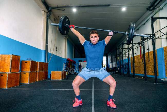 Potente atleta masculino sin levantar mano peso pesado durante el entrenamiento funcional cerca de equipos deportivos en el gimnasio - foto de stock