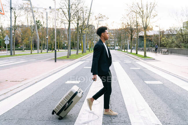 Side view of African American male with luggage walking along crosswalk in city — Stock Photo