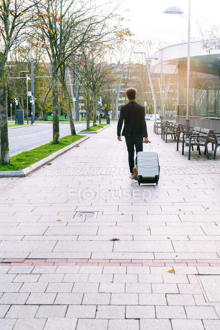 Back view of black male traveler in trendy outfit walking with suitcase in city and looking away — Stock Photo