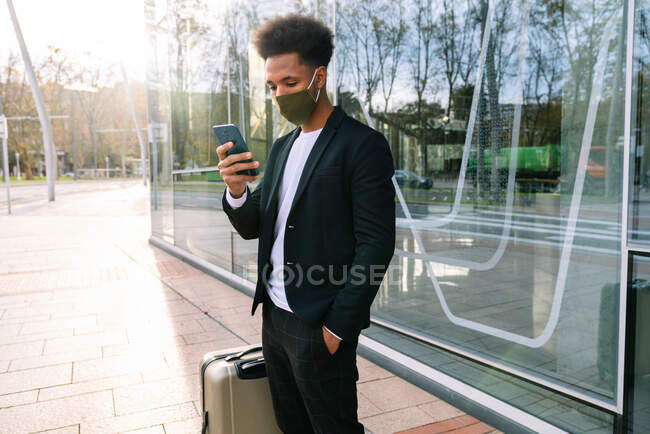 Afroamerikaner mit Schutzmaske steht neben Koffer auf der Straße und surft Smartphone, während er während der Coronavirus-Epidemie auf seine Abreise wartet — Stockfoto