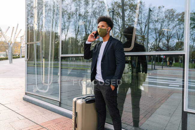 Афроамериканец в защитной маске стоит рядом с чемоданом на улице и разговаривает по смартфону, ожидая отъезда во время эпидемии коронавируса — стоковое фото