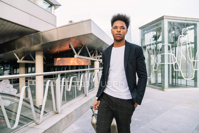 Serious black male traveler in trendy outfit walking with suitcase in city and looking away — Stock Photo