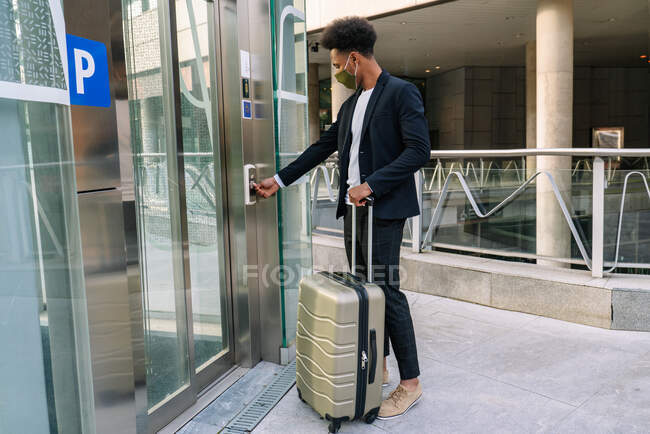 Вид сбоку афроамериканского туриста-мужчины с чемоданом и в защитной маске, нажимающей кнопку лифта в аэропорту во время путешествия во время пандемии коронавируса — стоковое фото