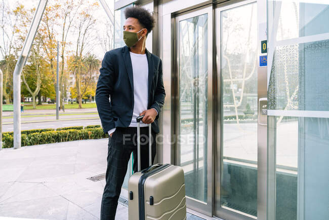 Turista masculino afroamericano con maleta y máscara protectora de pie cerca del ascensor en el aeropuerto mientras viaja durante la pandemia de coronavirus - foto de stock