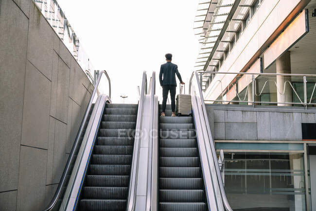 Низкий угол заднего вида анонимного мужчины-путешественника с чемоданом, стоящим на движущейся лестнице возле здания аэропорта — стоковое фото