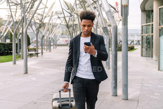 Serious African American male tourist walking with suitcase in airport and checking time of departure on smartphone — Stock Photo