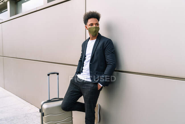 Афроамериканець чоловічої статі в захисній масці стоїть біля валізи і спирається на будівлю аеропорту в очікуванні польоту під час епідемії коронавірусу. — Stock Photo