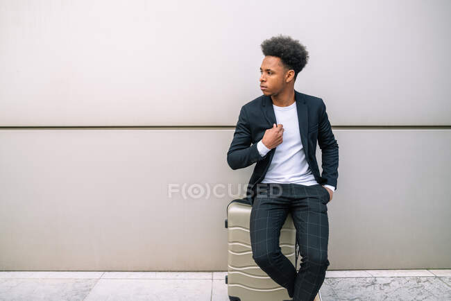 Мирный черный мужчина путешественник сидит на чемодане возле здания в аэропорту, ожидая отъезда и глядя в сторону — стоковое фото
