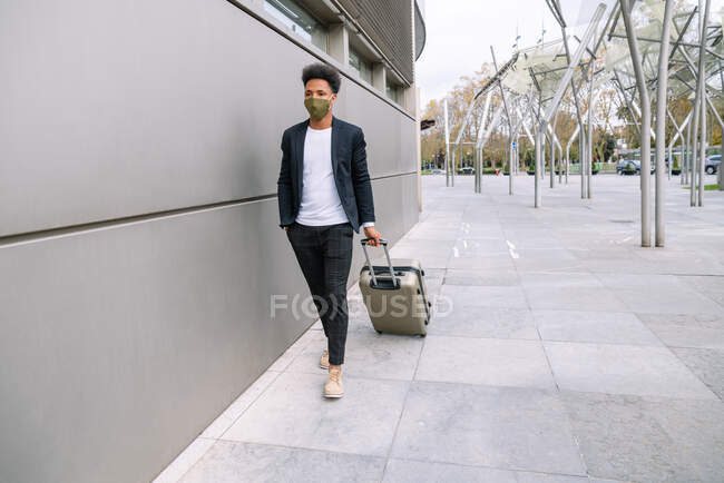 Viajero masculino negro serio en traje de moda y máscara caminando con maleta en la ciudad y mirando hacia otro lado - foto de stock