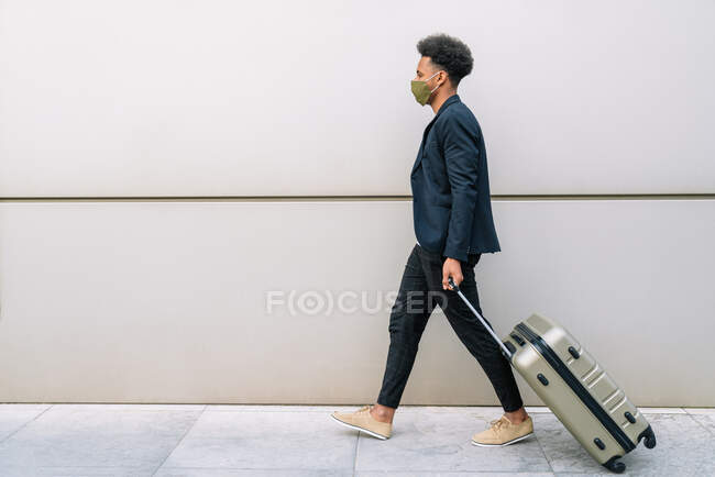 Side view black male traveler in trendy outfit and mask walking with suitcase in city and looking away — Stock Photo
