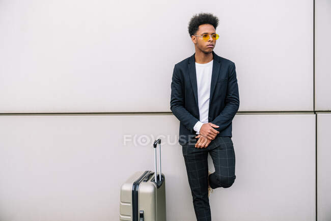 Voyageur homme noir en tenue tendance debout avec valise contre mur blanc en ville et détournant les yeux — Photo de stock