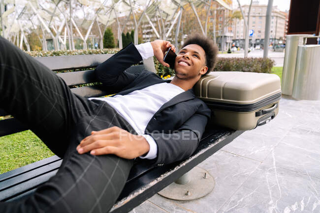 Веселый афроамериканец-путешественник лежит на чемодане на скамейке и говорит по мобильному телефону — стоковое фото