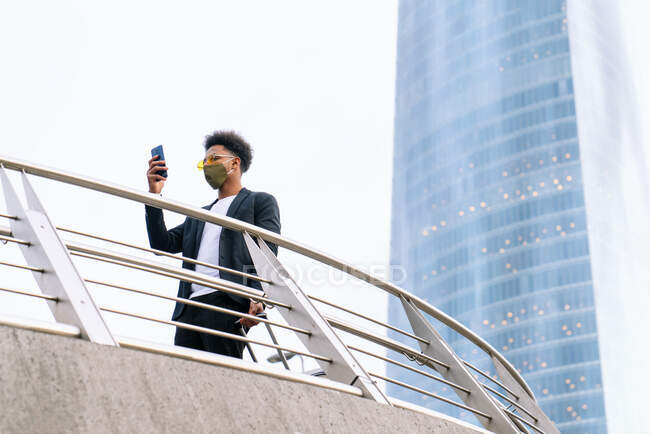 Von unten afroamerikanische männliche Reisende in Schutzmaske und mit Koffer, der in Megapolis steht und während der Reise mit dem Coronavirus im Smartphone surft — Stockfoto
