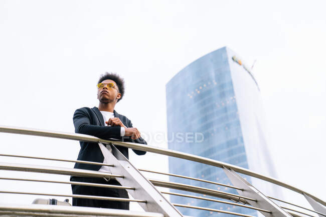 De baixo de confiante afro-americano masculino inclinado sobre trilhos enquanto de pé no centro da cidade e olhando para longe — Fotografia de Stock