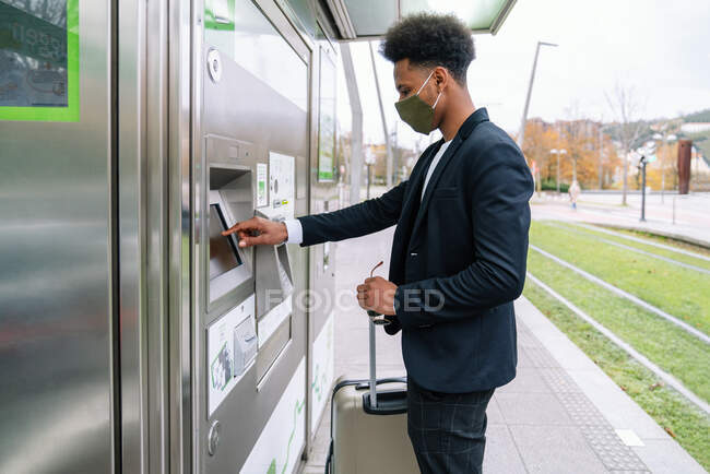 Vista lateral de un hombre afroamericano enmascarado usando una máquina expendedora de billetes mientras está parado en la estación de tren con la maleta y viajando durante el coronavirus - foto de stock