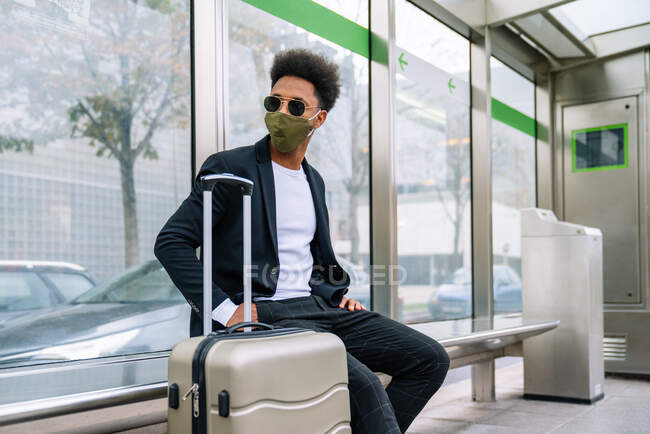 Side view of traveling African American male in protective mask sitting on bench with suitcase and looking away — Stock Photo