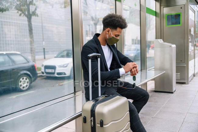 Seitenansicht eines reisenden Afroamerikaners in Schutzmaske, der mit einem Koffer auf einer Bank sitzt und die Ankunftszeit des Zuges überprüft — Stockfoto