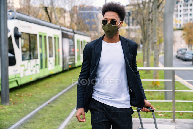 Preto viajante masculino com mala e máscara protetora em pé no planform na estação ferroviária e esperando por trem durante coronavírus — Fotografia de Stock