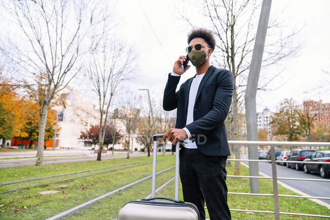 Black male traveler with suitcase and in protective mask talking phone while waiting for train during coronavirus — Stock Photo