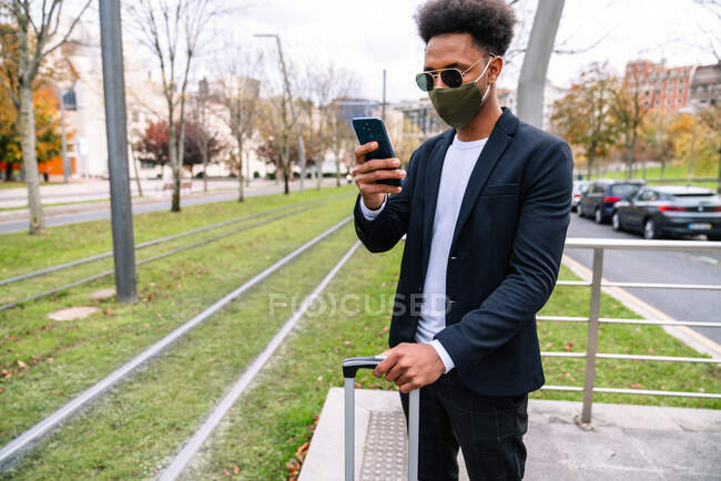 Viaggiatore maschio nero con valigia e in maschera protettiva telefono di navigazione in attesa di treno durante il coronavirus — Foto stock