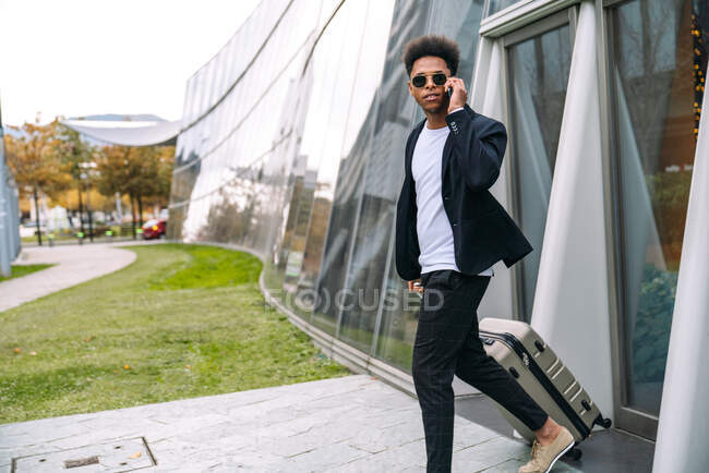 Seitenansicht eines schwarzen männlichen Touristen mit Koffer, der in der Nähe eines gläsernen städtischen Gebäudes unterwegs ist, während er am Handy spricht und wegschaut — Stockfoto