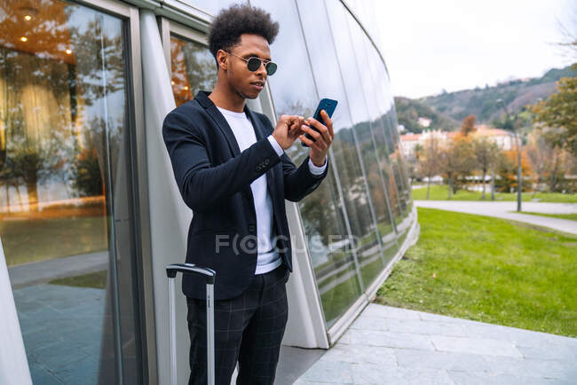 Вид сбоку чернокожего туриста-мужчины с чемоданом, идущего рядом со стеклянным городским зданием во время просмотра мобильного телефона — стоковое фото