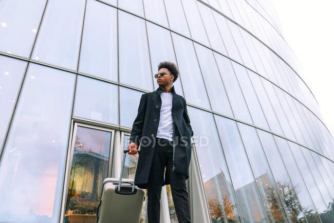 De baixo de viajante masculino preto na roupa da moda em pé com mala contra o edifício de vidro moderno na cidade e olhando para longe — Fotografia de Stock