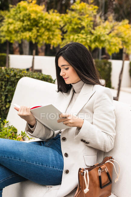 Вид сбоку женщины-предпринимателя, сидящей на скамейке в парке и проверяющей расписание в блокноте — стоковое фото