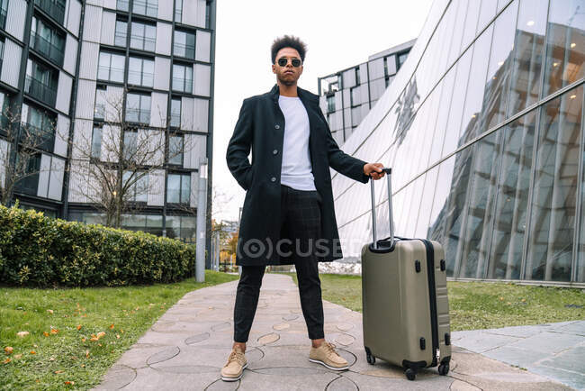 From below of black male traveler in trendy outfit standing with suitcase against modern glass building in city and looking away — Stock Photo