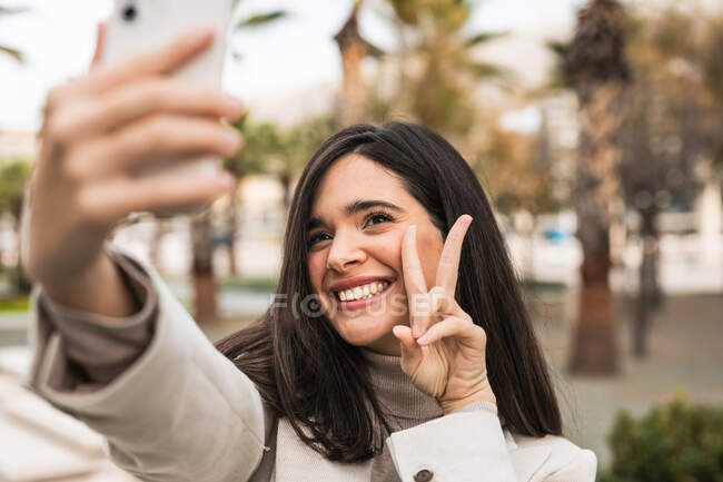 Улыбающаяся очаровательная женщина делает селфи на смартфоне, стоя на улице с пальмами — стоковое фото