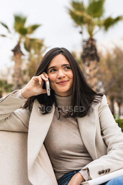 Alegre empresaria sentada en el banco de la ciudad y hablando por teléfono móvil mientras sonríe y mira hacia otro lado - foto de stock