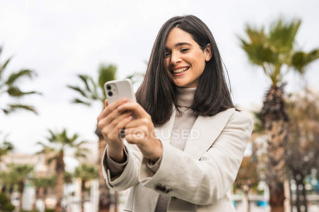 Charmant lächelnde Frau macht Selfie mit dem Smartphone, während sie auf der Straße mit Palmen steht — Stockfoto