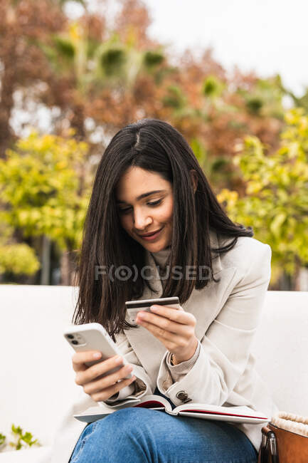 Mulher sorridente sentada no banco e fazendo compra com cartão de plástico via smartphone durante compras on-line — Fotografia de Stock